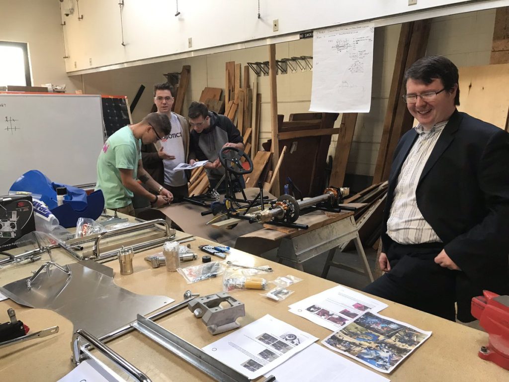 two male students in the background working on a go-kart, student in the foreground smiling at his work