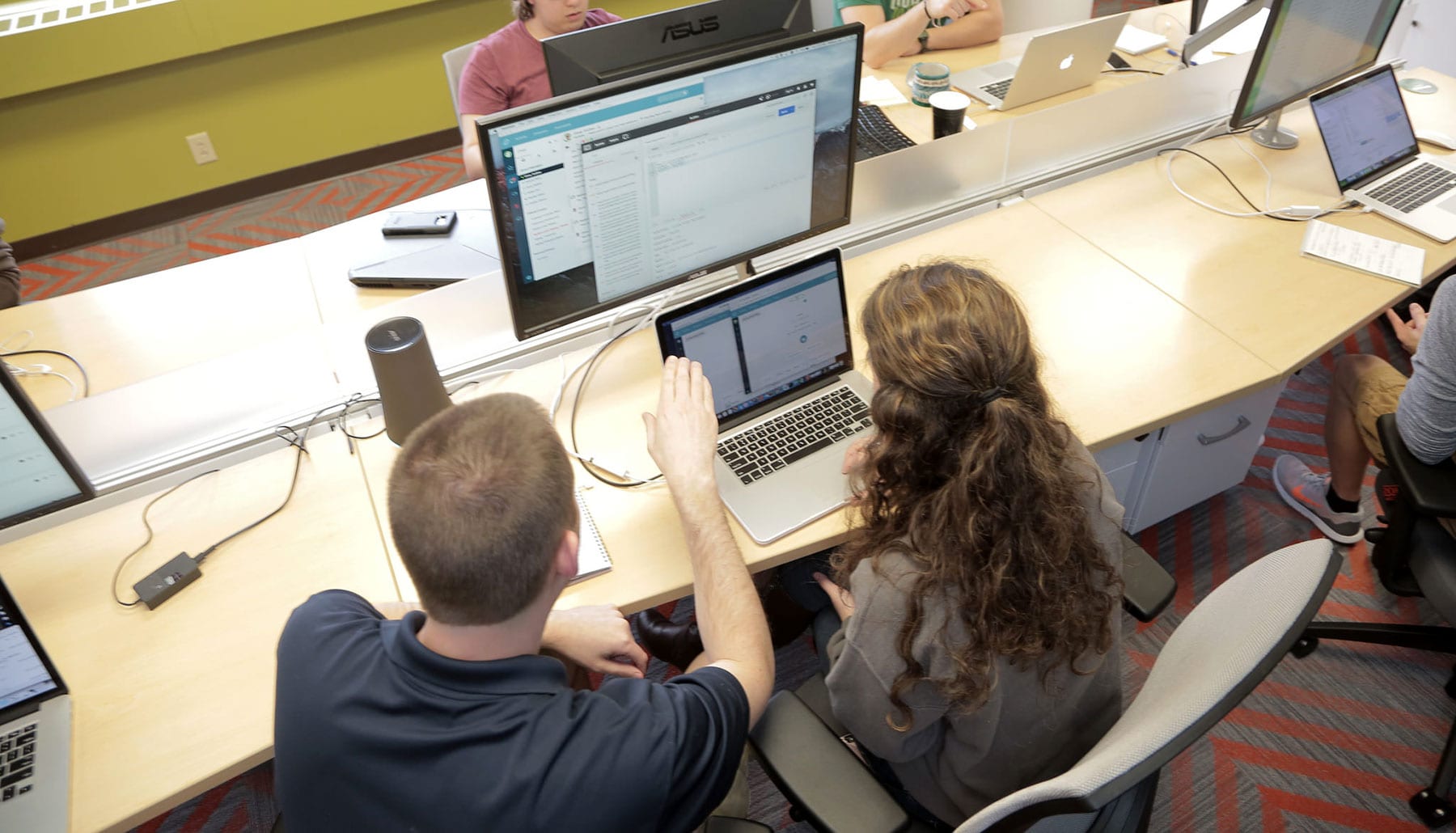 A man and young woman are viewed from above and behind working on two computers.